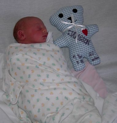 A newborn picture of Jameson, wrapped in a receiving blanket and lying next to a stitched bear with his name printed on it