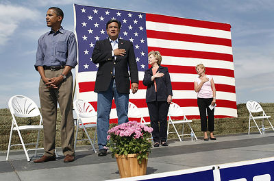 Barack Obama, Bill Richardson, Hillary Clinton, and Ruth Harkin are standing on a stage with a large American flag as the backdrop.  All but Obama have their right hands over their hearts; he has his hands clasped in front of him.