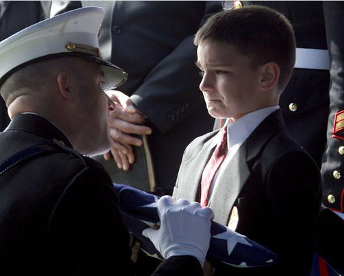 A boy is standing in a suit, with a sad but determined face, as an officer hands him a folded flag